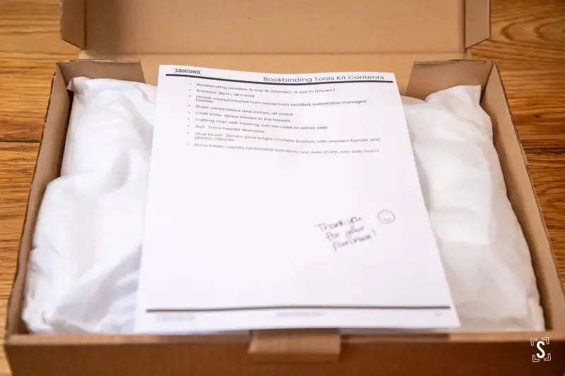 Open cardboard box with tissue paper and document for Kit Bookbinding Tools contents
