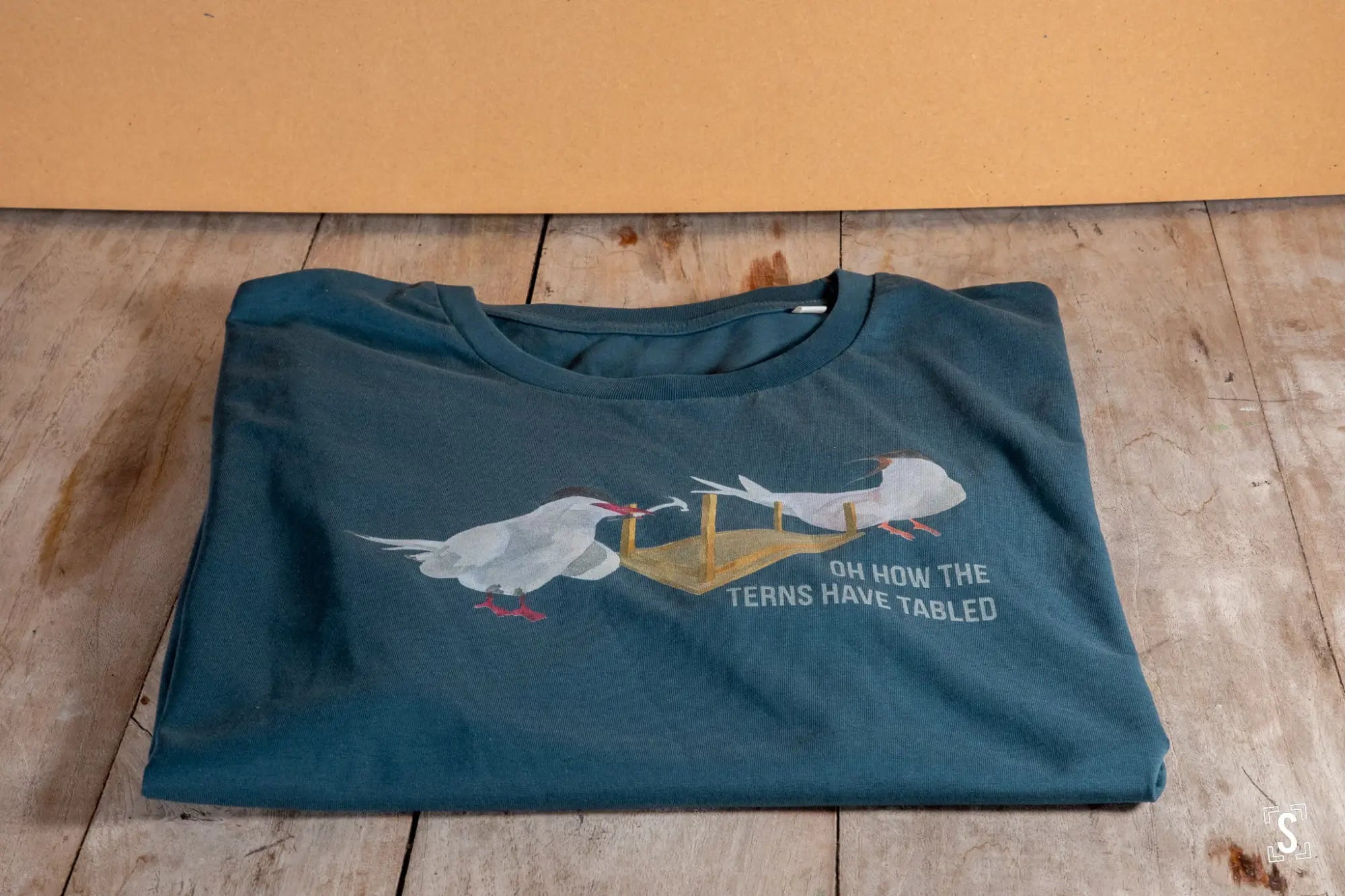 Folded teal t-shirt with a goose carrying a table printed on it.