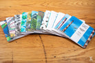 Colorful travel brochures fan out on a wooden surface beside a Handmade Notebook with chain stitch binding