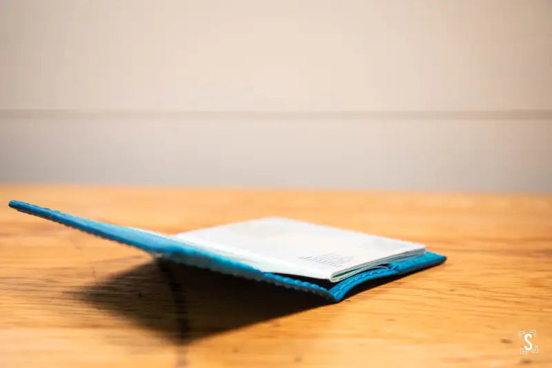 Open book with blue cover on wooden surface next to Leathercraft Workshop passport holder